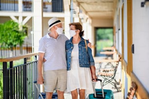 Senior couple with face masks and luggage outside apartment on holiday, back to normal concept.
