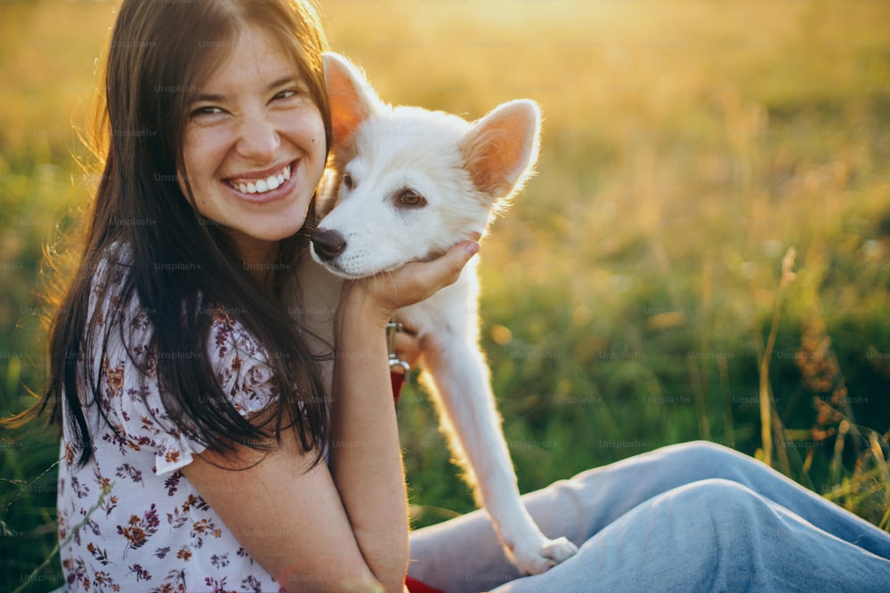 Mulher feliz abraçando filhote de cachorro branco bonito no prado de verão na luz do pôr do sol. Autêntico momento bonito. Menina elegante sorrindo e relaxando com seu filhote adorável em um piquenique