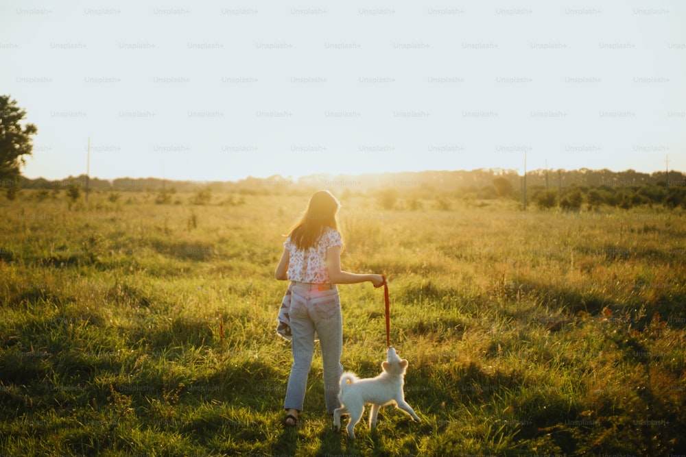 Elegante chica hipster que camina con un lindo cachorro blanco en el prado de verano. Mujer joven en un paseo con su perrito en la cálida luz del sol. Concepto de adopción, amigo leal
