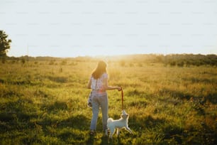 Stylish hipster girl walking with cute white puppy in summer meadow. Young woman on a walk with her doggy in warm sunshine light. Adoption concept, loyal friend