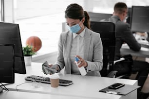 Businesswoman with medical mask disinfecting desk in the office.
