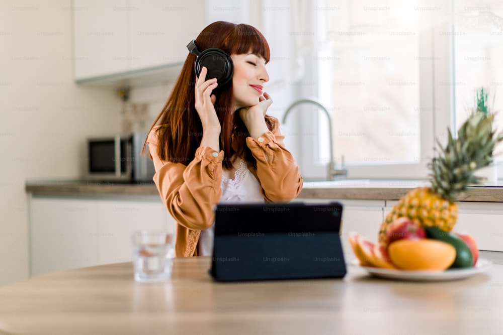 Young pretty girl in headphones is listening to music while working on tablet and having healthy breakfast at home. Lifestyle portrait of modern woman starting day with fruits and music.