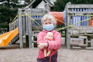 Sad Caucasian girl in face mask with baby toy on closed playground outdoor. Kids play area locked with yellow caution tape in Toronto Canada. Coronavirus social distance quarantine.