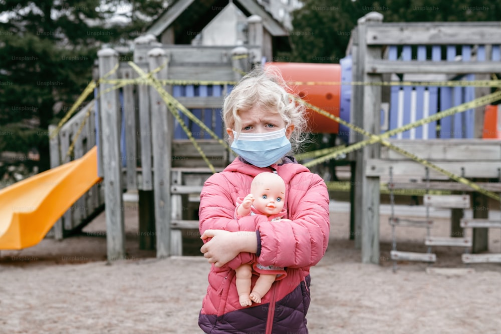 Sad Caucasian girl in face mask with baby toy on closed playground outdoor. Kids play area locked with yellow caution tape in Toronto Canada. Coronavirus social distance quarantine.