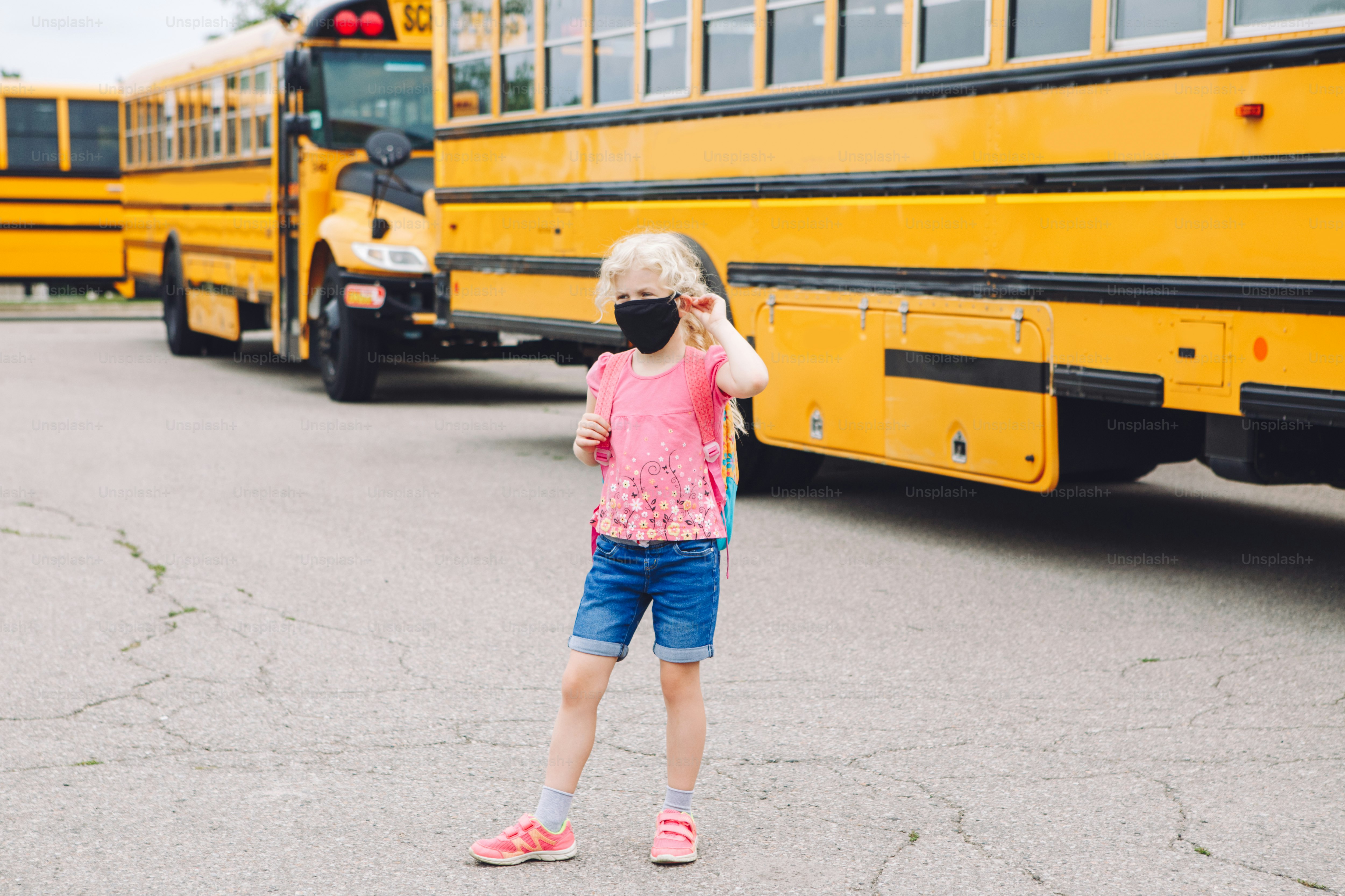 Happy Caucasian girl student wearing face mask near yellow bus. Kid with personal protective equipment on face. Education and back to school in September. New normal during coronavirus.