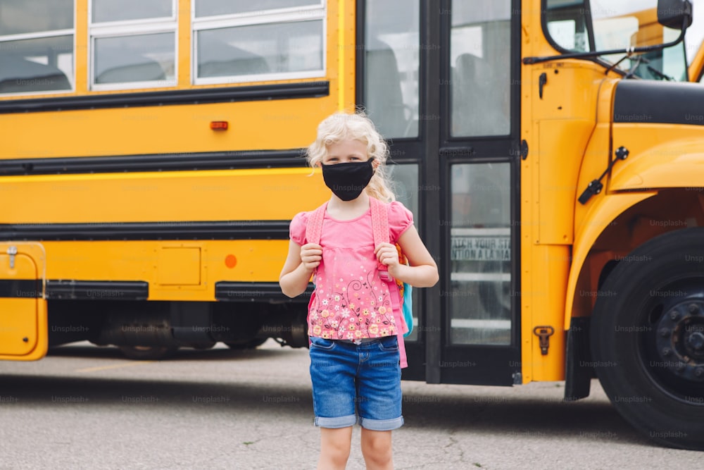 Happy Caucasian girl student wearing face mask near yellow bus. Kid with personal protective equipment on face. Education and back to school in September. New normal during coronavirus.