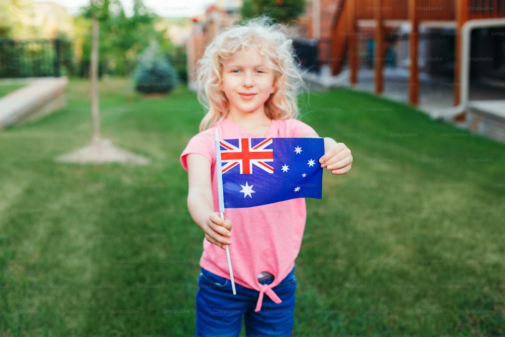 Adorable cute happy Caucasian girl holding Australian flag. Smiling child waving Australia flag. Kid citizen celebrating Australia Day holiday in January outdoor.