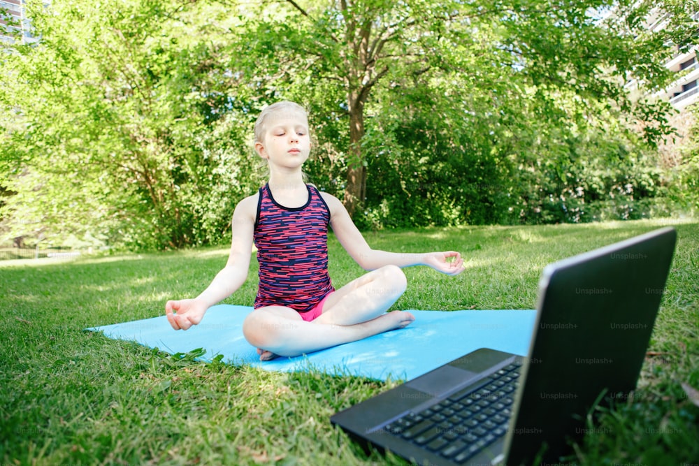 Niña practicando yoga al aire libre en línea. Video entrenamiento deportivo en Internet. Niño aprendiendo entrenamiento físico con computadora portátil. Nueva normalidad. Distancia social y educación en coronavirus. Concepto de alivio del estrés.