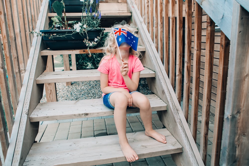 Adorable cute happy Caucasian girl holding Australian flag. Funny child kid covering her face with Australia flag. Little citizen celebrating Australia Day holiday in January outdoor.