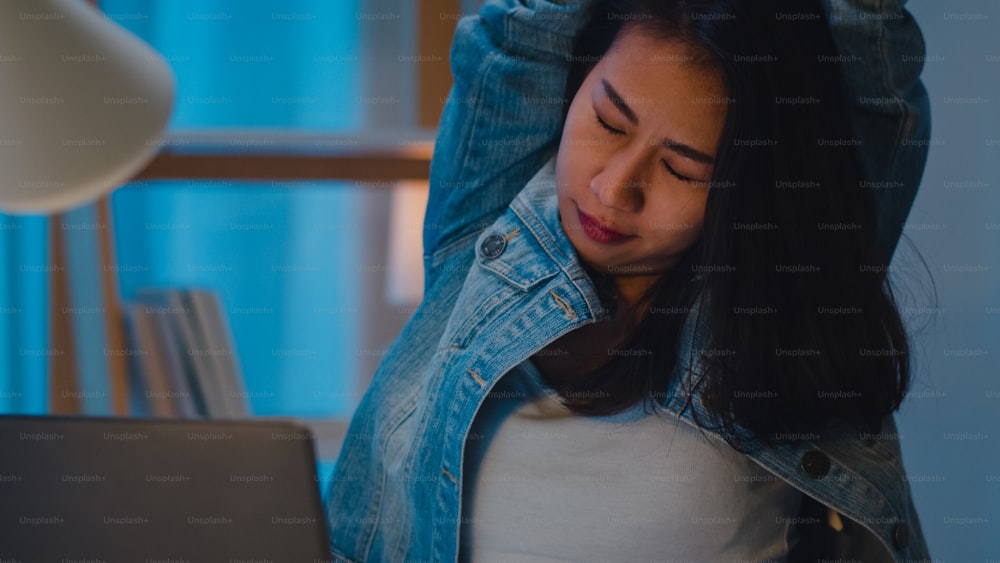 Millennial young Chinese businesswoman working late night stretching on her chair, looking in laptop computer in living room at modern home. Asia people occupational burnout syndrome concept.