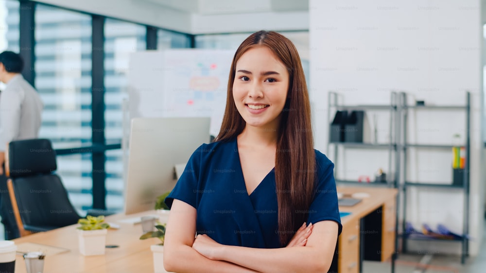 Portrait of successful beautiful executive businesswoman smart casual wear looking at camera and smile, arms crossed in modern office workplace. Young Asia lady standing in contemporary meeting room.