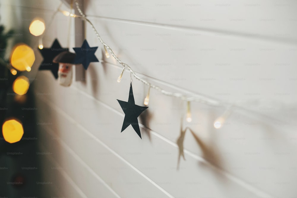 Stylish christmas stars and golden lights bokeh on background of white wall. Decorating festive scandinavian room for winter holidays. Merry Christmas and Happy Holidays. Space for text