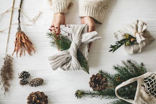 Manos que sostienen un elegante regalo de Navidad envuelto en tela de lino con rama de abeto verde y bayas rojas sobre una mesa blanca rústica con piñas, plana. Navidad sin residuos, vacaciones sin plástico