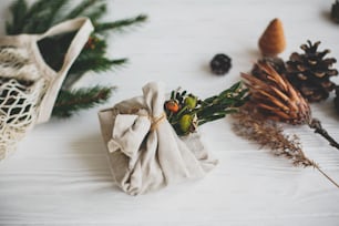 Stylish christmas gift wrapped in linen fabric, decorated with natural green branch on white rustic table background with pine cones and herbs. Zero waste winter holidays.