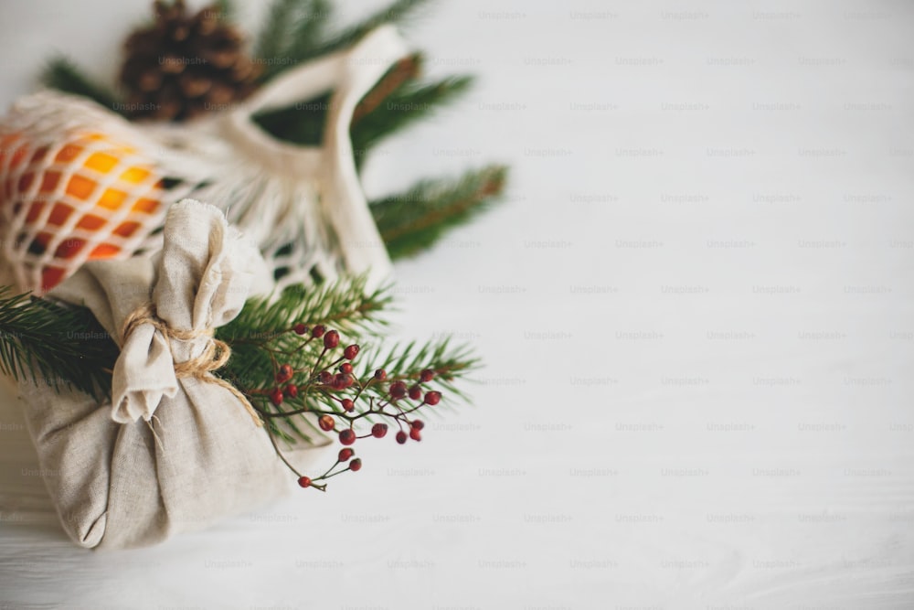 Zero waste christmas holidays. Stylish christmas gift wrapped in linen fabric with natural green branch and red berries on background of reusable cotton bag with oranges on white  table.