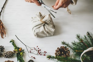 Hands wrapping stylish christmas gift in linen fabric on white rustic table with fir, pine cones, scissors, twine. Female preparing plastic free christmas present, zero waste holidays.