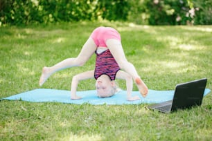 Girl child doing sport workout outdoor online. Video yoga stretching on Internet in park. Kid learning fitness gymnastics on backyard with laptop. New normal. Social distance at coronavirus.