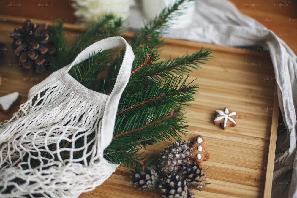 Zero Waste Weihnachtsferien. Wiederverwendbare Einkaufstasche mit grünen Fichtenzweigen, Lebkuchen-Weihnachtsplätzchen und Tannenzapfen auf rustikalem Holzhintergrund. Plastikfreie Winterdekoration