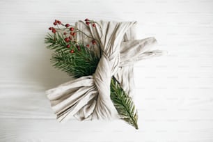 Stylish christmas gift, flat lay. Present wrapped in linen fabric and decorated with natural fir branch and red berries on white rustic table background. Zero waste winter holidays.
