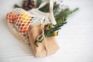 Zero Waste Weihnachtsferien. Einfache Weihnachtsgeschenkbox eingewickelt in Bastelpapier mit naturgrünem Zweig auf dem Hintergrund einer wiederverwendbaren Baumwolltasche mit Orangen und Tannenzweigen auf weißem Tisch.