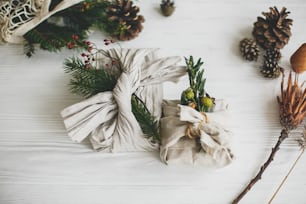 Stylish christmas gifts wrapped in linen fabric, decorated with natural green branch on white rustic table background with pine cones and herbs. Zero waste winter holidays.