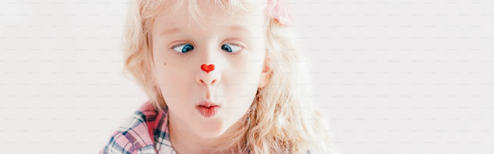 Niño bizco mirando la pegatina de corazón rojo en la nariz. Divertida e hilarante niña blanca caucásica linda adorable con ojos azules. Concepto de vacaciones del día de San Valentín. Encabezado de banner web para sitio web.