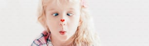 Cross-eyed kid looking at red heart sticker on nose. Funny hilarious white Caucasian cute adorable child girl with blue eyes. Valentine Day holiday concept. Web banner header for website.