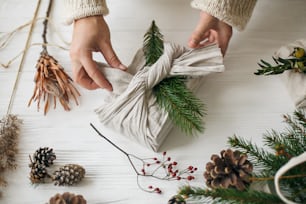 Florist preparing zero waste Christmas gift. Plastic free holidays. Hands decorating stylish christmas gift in linen fabric with green fir branch on white rustic table with pine cones and berries.