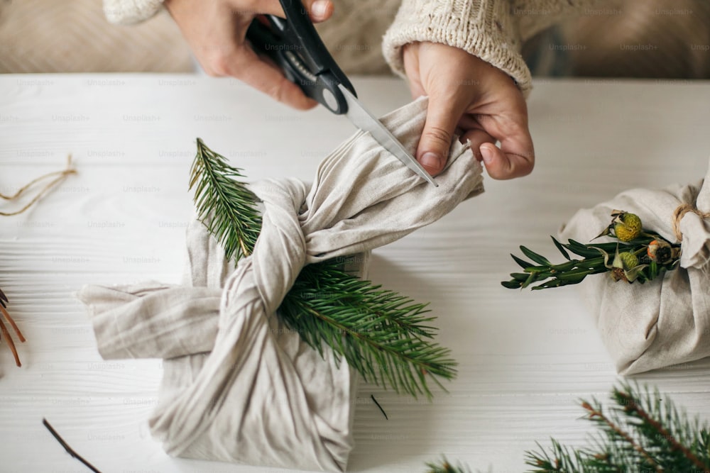 Hands wrapping gift in linen fabric with natural green branch on background of white rustic table. Florist preparing present, holding scissors. Zero waste stylish christmas gift.