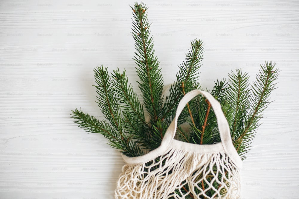 Net shopping bag with winter decorations, zero waste holidays. Sustainable lifestyle. Reusable cotton bag with green spruce branches on white rustic background.