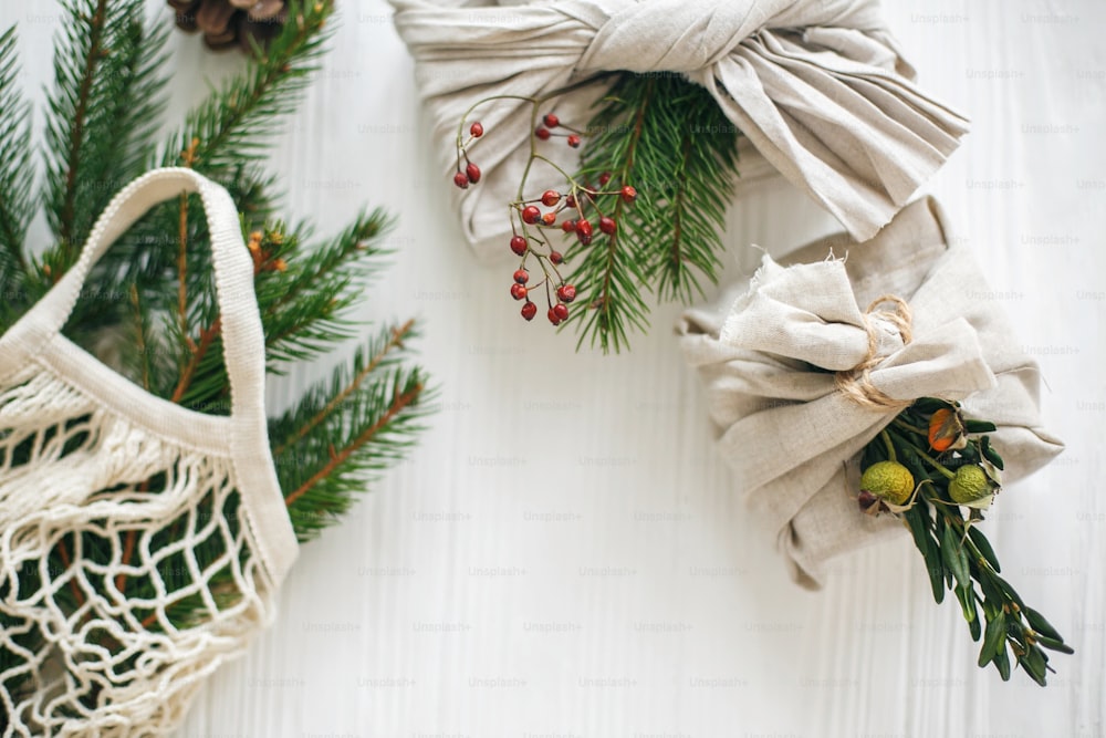 제로 폐기물 크리스마스 휴일. 녹색 가지가 있는 린넨 패브릭으로 싸인 세련된 크리스마스 선물과 소박한 나무 배경에 녹색 가문비나무가 있는 재사용 가능한 쇼핑백.
