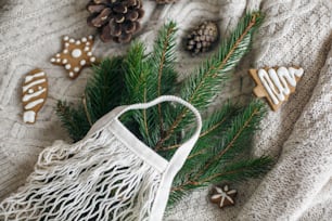 Reusable shopping bag and wooden winter decorations, plastic free holidays. Net cotton bag with green spruce branches, gingerbread christmas cookies and pine cones on white knitted sweater.