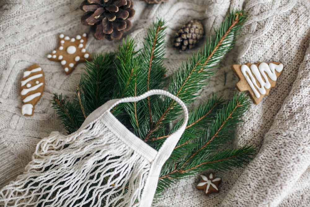 再利用可能なショッピングバッグと木製の冬の装飾、プラスチックフリーの休日。白いニットセーターに緑のトウヒの枝、ジンジャーブレッドのクリスマスクッキー、松ぼっくりのネットコットンバッグ。
