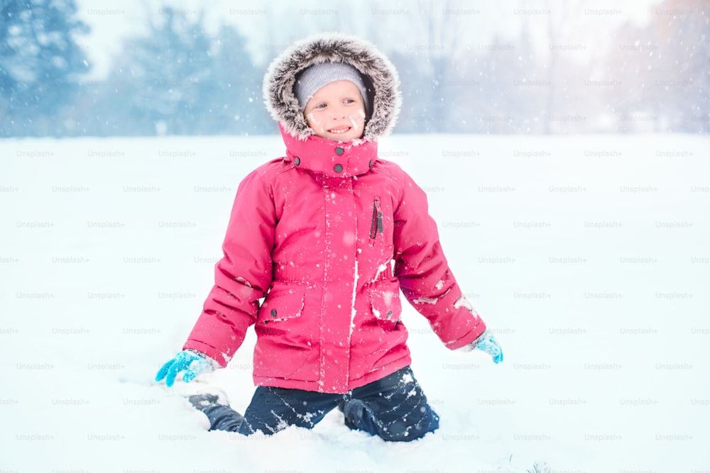 Cute adorable funny Caucasian smiling girl child in warm clothes red pink jacket playing with snow. Kid having fun during cold winter snowy day at snowfall. Kids outdoor seasonal activity.
