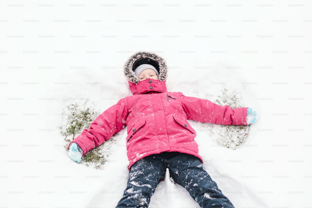 Cute adorable funny Caucasian girl child in warm clothes pink jacket making snow angel. Kid lying on ground during cold winter snowy day. Kids outdoor seasonal activity. View from top above.