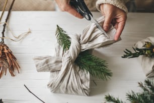 Zero Waste stilvolles Weihnachtsgeschenk. Hände wickeln Geschenk in Leinenstoff mit natürlichem grünen Zweig auf dem Hintergrund des weißen rustikalen Tisches. Florist bereitet Geschenk vor, hält eine Schere