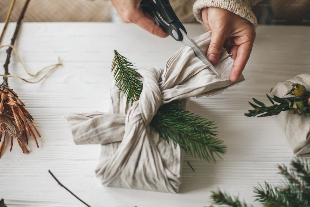 Zero waste stylish christmas gift. Hands wrapping gift in linen fabric with natural green branch on background of white rustic table. Florist preparing present, holding scissors