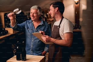 Cheerful young man standing with a clipboard in a cellar and looking at a glass of red wine in hand of a positive colleague