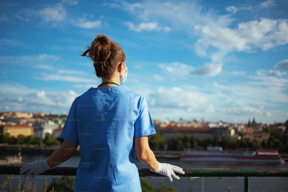 Pandemia di coronavirus. Visto da dietro, medico moderno, donna in camice con maschera medica e guanti di gomma, che guarda in lontananza fuori contro il cielo.