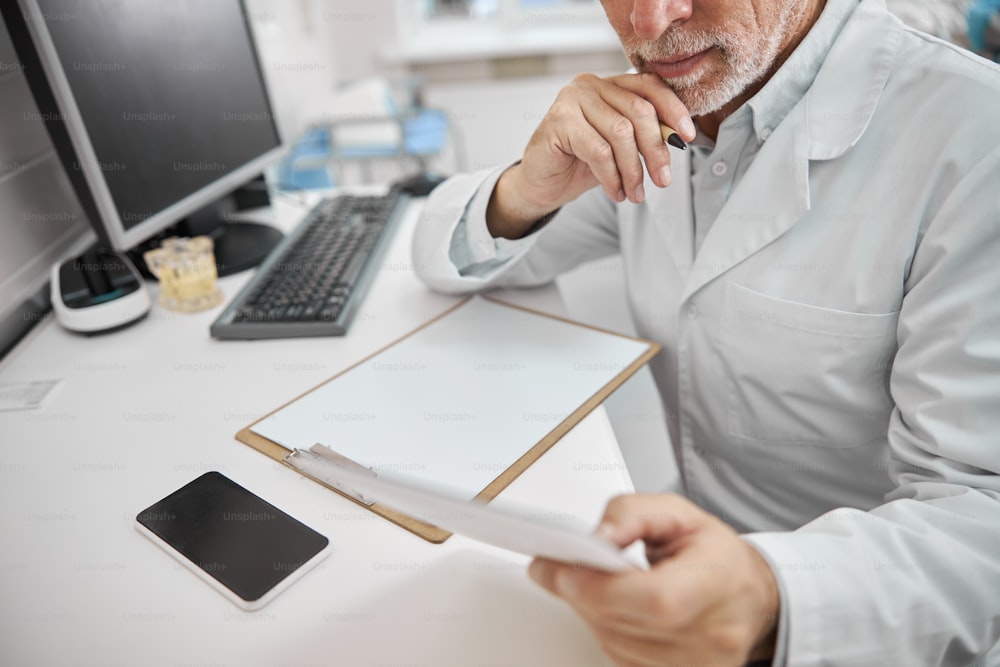 Cropped photo of a professional elderly doctor looking at the tablet while sitting at his office desk