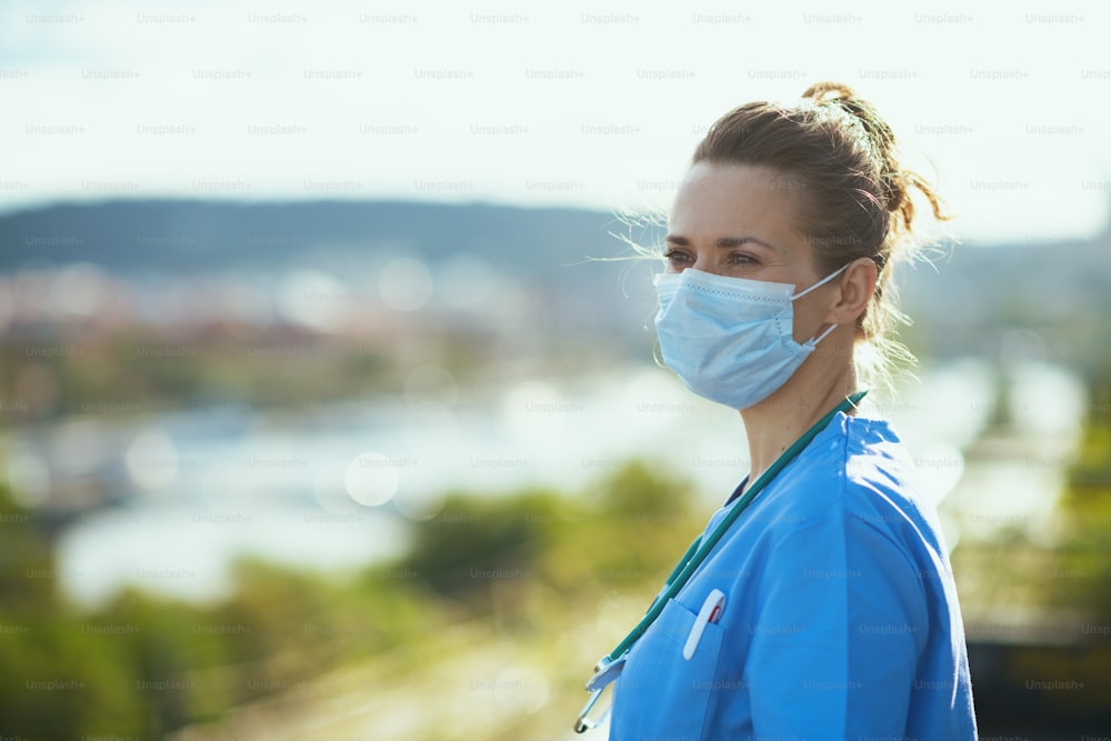 코비드-19 팬데믹. 청진기와 의료용 마스크를 쓴 제복을 입은 잠겨 있는 현대 의사 여성이 도시 거리의 야외에서 먼 거리를 바라보고 있습니다.