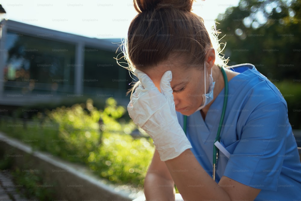 Pandemia di coronavirus. donna medica moderna stressata in uniforme con stetoscopio e maschera medica seduta all'aperto vicino all'ospedale.