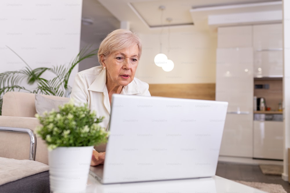 Ritratto di donna anziana seduta al tavolo a casa e che lavora sul suo laptop. Signora anziana che naviga in rete da casa mentre si siede sul suo divano e utilizza il computer portatile