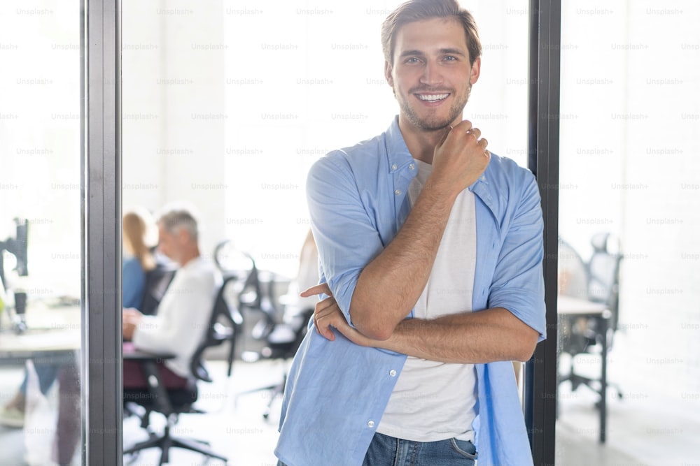 Happy businessman standing in the office with coworkers in the background working by the desk