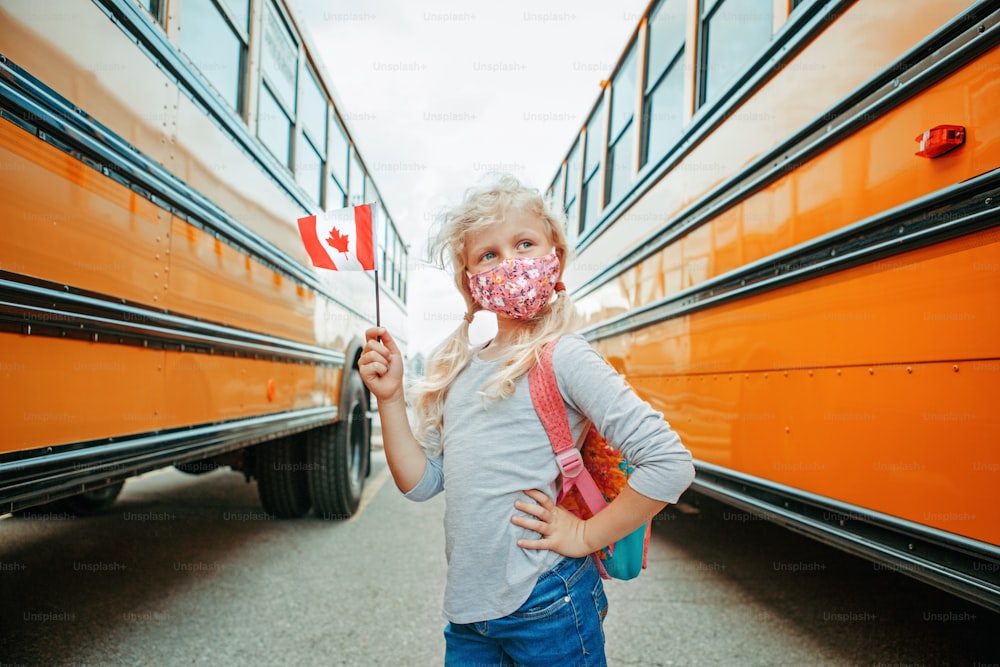Studentessa caucasica con maschera facciale che tiene bandiera canadese. Bambino studente vicino allo scuolabus giallo in Canada. Istruzione e ritorno a scuola a settembre. Nuova normalità durante il coronavirus.