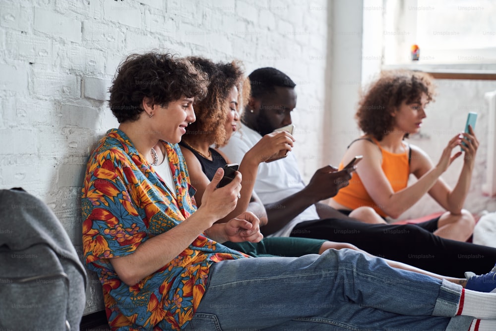Side view of smiling friends holding mobile phones while sitting on the floor near white wall