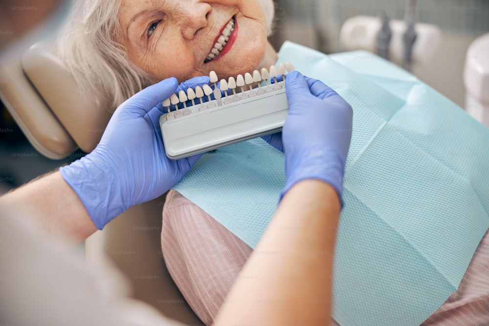 Retrato em close up do médico escolhendo a cor de um membro artificial dentário com paciente do sexo feminino na clínica odontológica