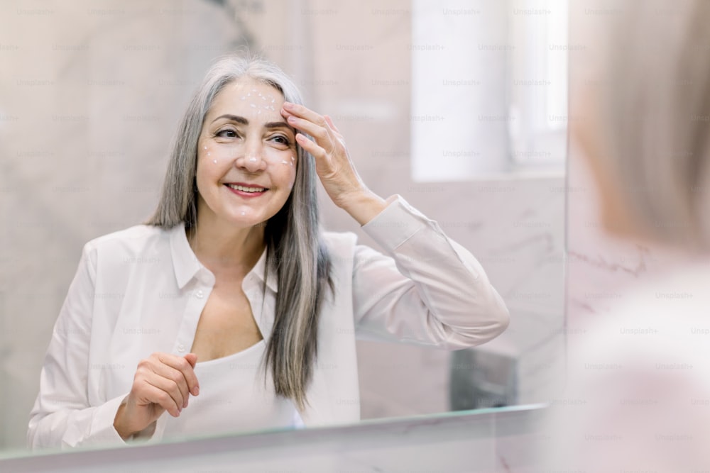 Pretty senior woman with long gray hair, wearing white shirt, looking at her face in the bathroom mirror, and applying anti-wrinkle cream or cosmetic lotion. Skin care, antiaging treatment.