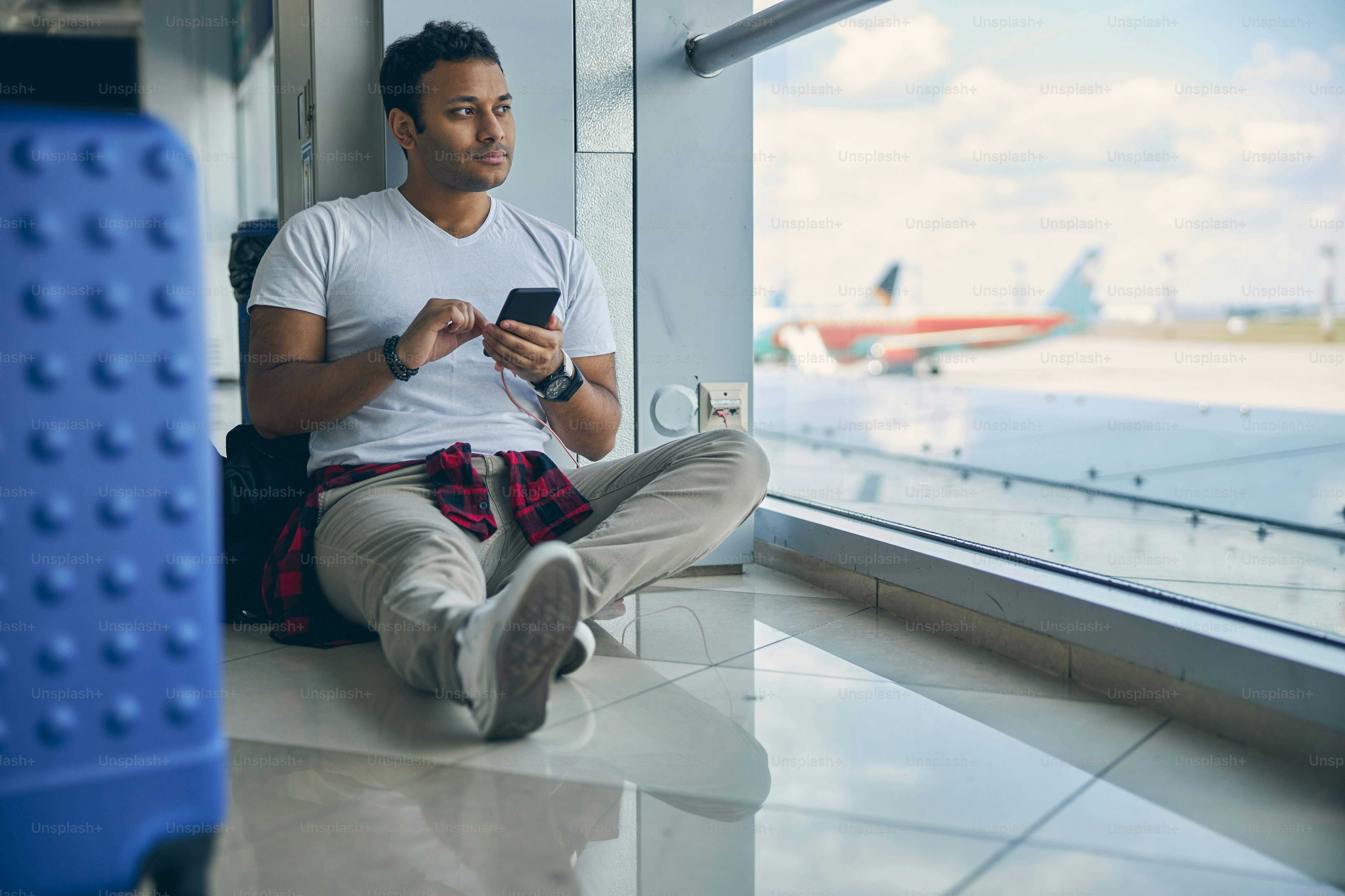 Full length portrait of intelligent man in casual clothes typing message on mobile phone while sitting on the floor with plane on background