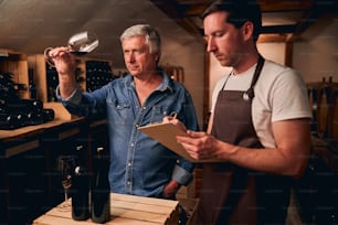 Calm Caucasian man standing in a winery cellar with his younger colleague and looking at the wine while tilting a glass